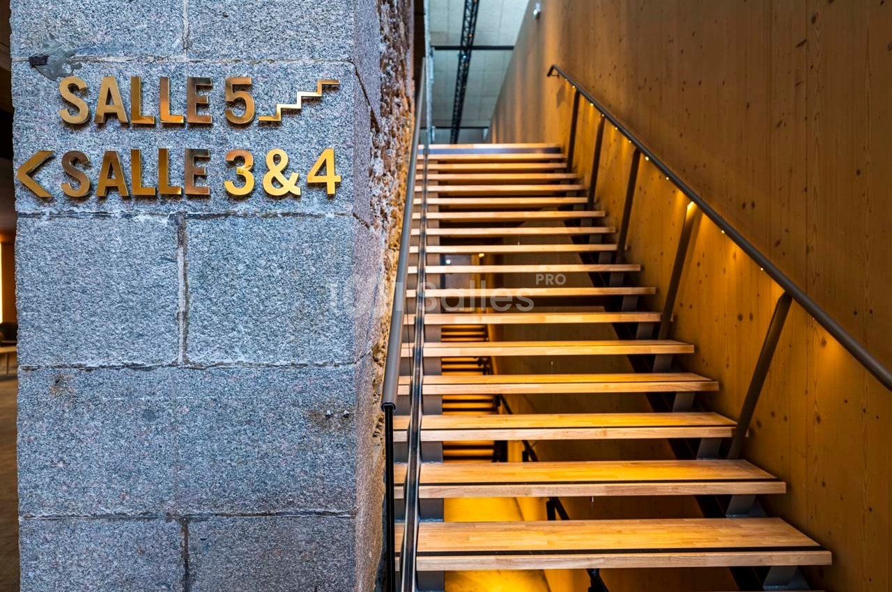 Escalier en bois éclairé menant aux salles 3, 4 et 5, avec des panneaux directionnels sur un mur en pierre.