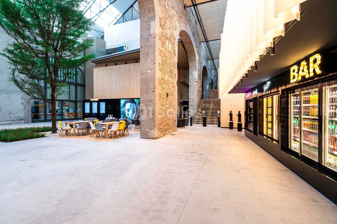 Espace intérieur moderne avec tables, arbre, arc en pierre, distributeurs de boissons et enseigne lumineuse ’BAR’.