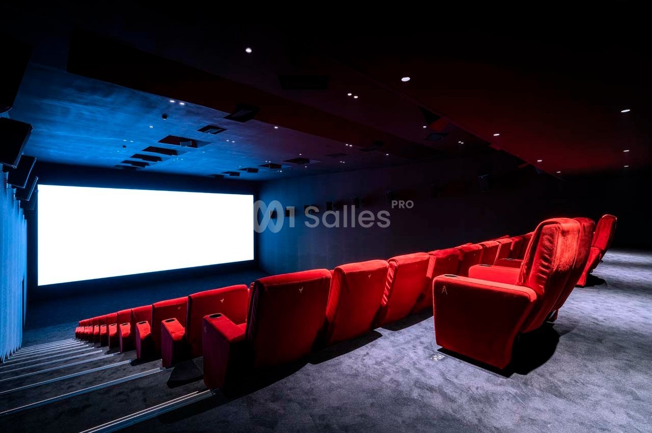 Salle de cinéma vide avec des rangées de fauteuils rouges et un grand écran blanc allumé.