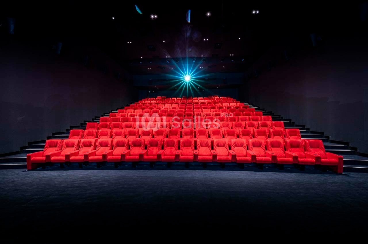 Salle de cinéma vide avec des rangées de sièges rouges et un projecteur allumé au fond.