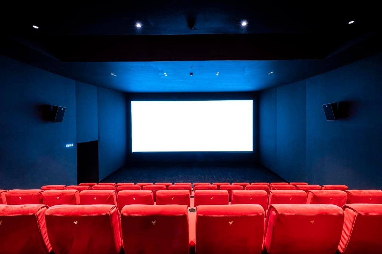 Salle de cinéma vide avec des rangées de fauteuils rouges face à un grand écran blanc allumé.