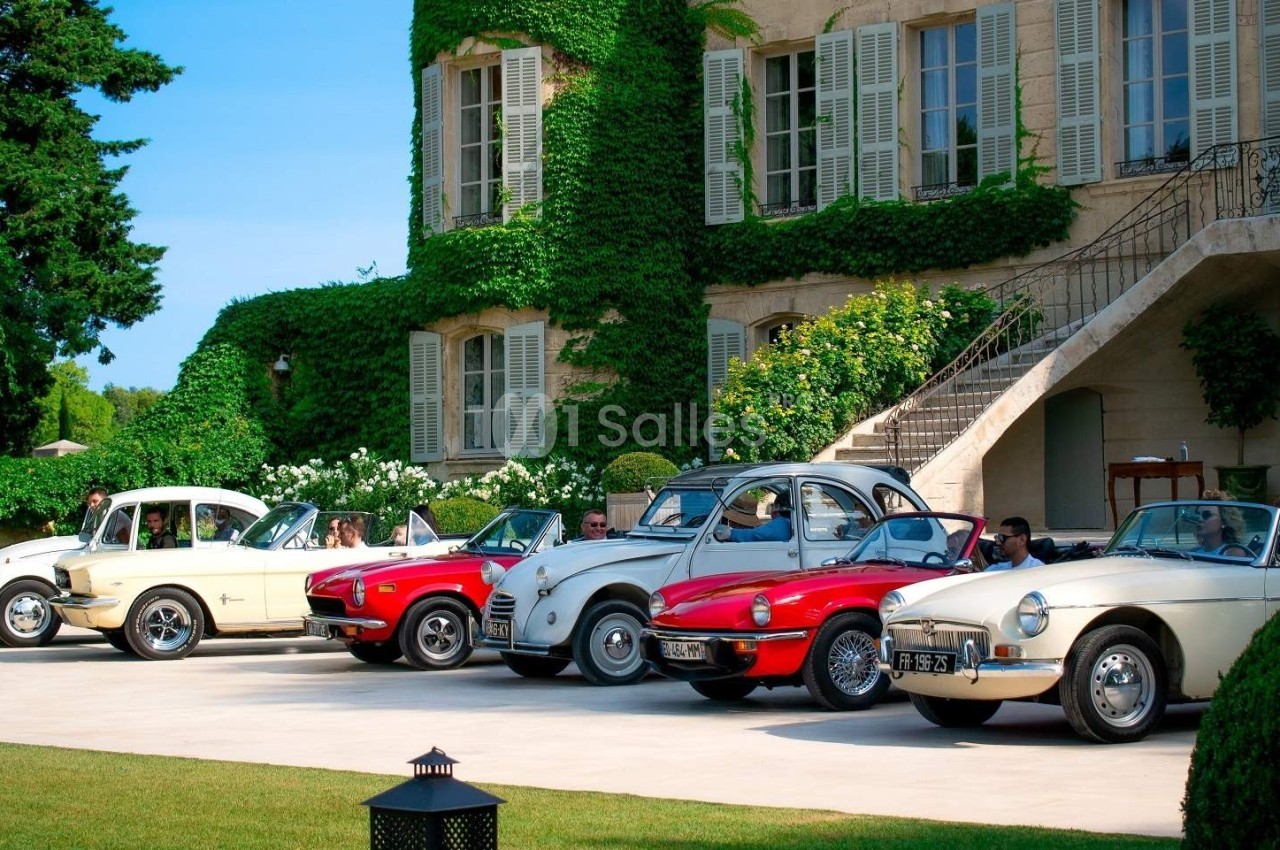 Alignement de voitures anciennes colorées devant une grande maison en pierre couverte de lierre.