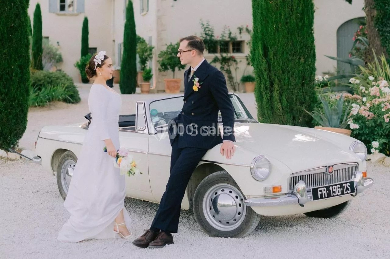 Un couple en tenue de mariage pose devant une voiture ancienne blanche dans une cour bordée de cyprès.