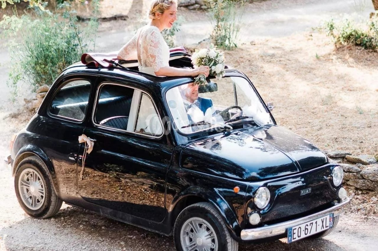 Une mariée souriante sort d'une petite voiture noire vintage, tenant un bouquet de fleurs blanches.