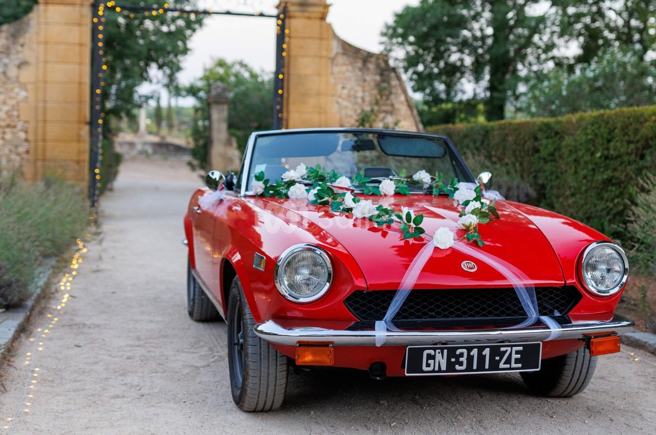 Voiture décapotable rouge décorée de fleurs blanches et rubans, stationnée devant une entrée en pierre.