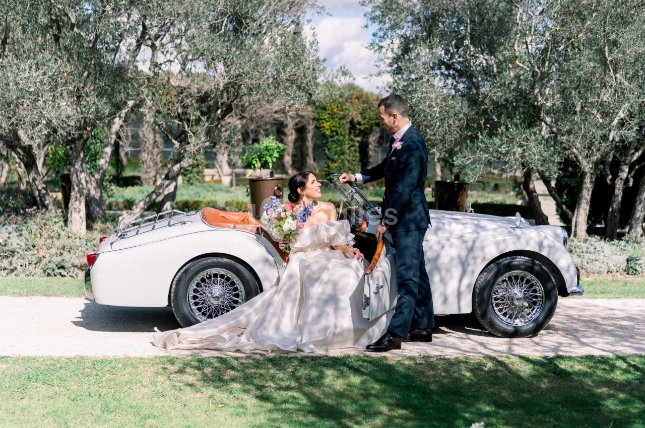 Un couple élégant pose près d'une voiture ancienne blanche dans un jardin bordé d'oliviers.