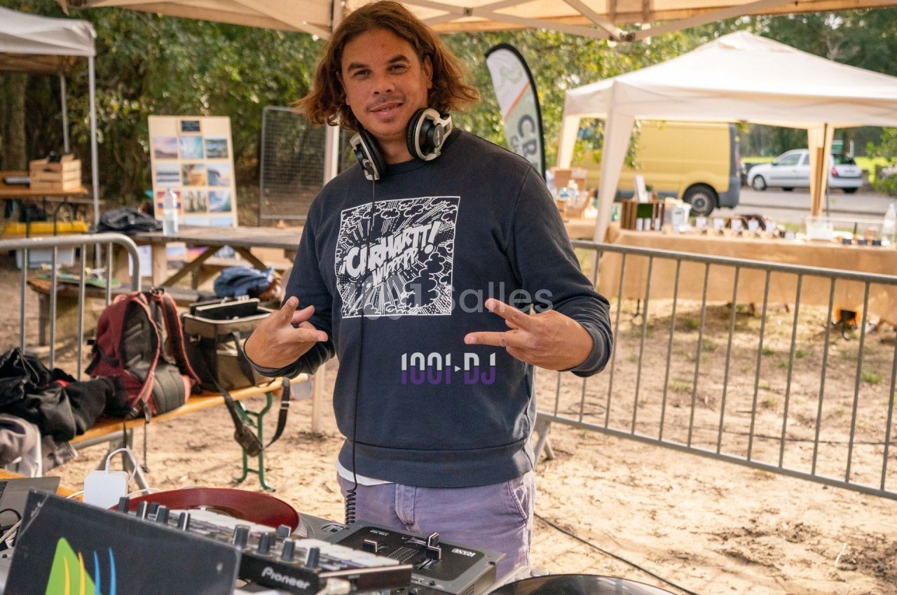 Un DJ souriant devant sa table de mixage dans un espace extérieur avec des stands en arrière-plan.