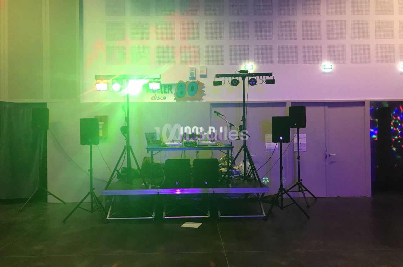 Scène de DJ avec éclairage coloré, platines et enceintes dans une salle aux murs clairs.