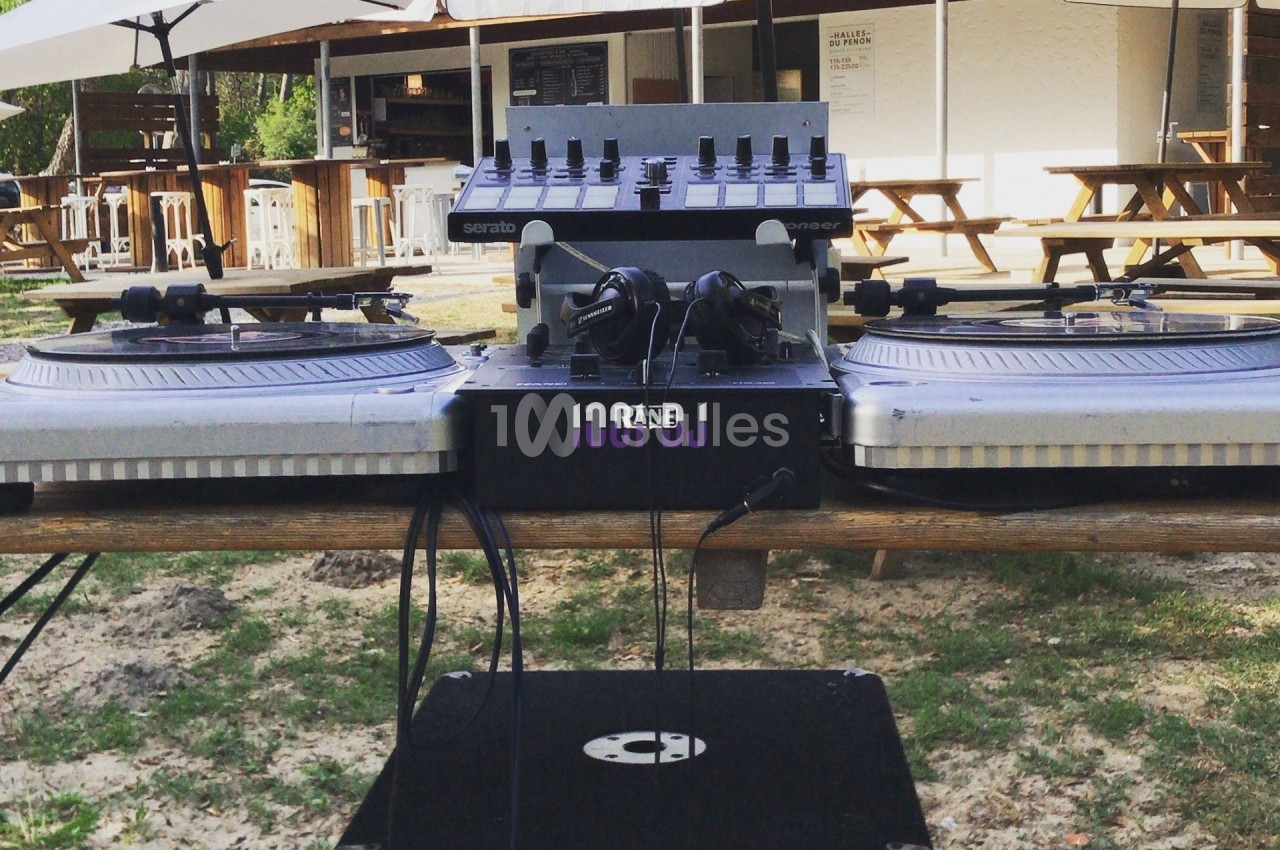 Deux platines vinyles et une table de mixage installées en extérieur sous des parasols, avec des tables en arrière-plan.