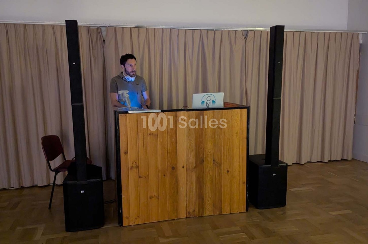 Un homme debout derrière un pupitre en bois avec un ordinateur portable, dans une salle avec rideaux beiges.