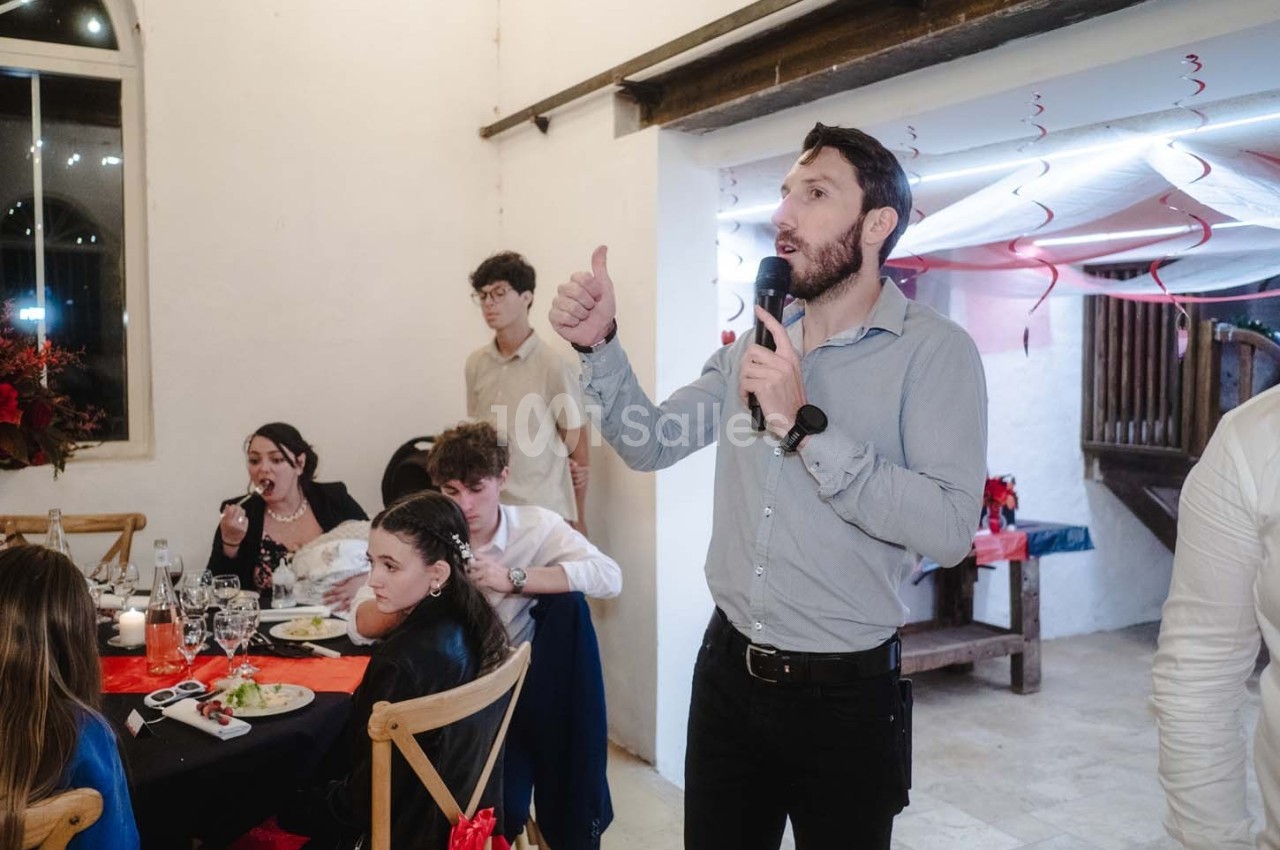 Un homme debout avec un micro s'adresse à des convives assis à une table lors d'un événement festif.