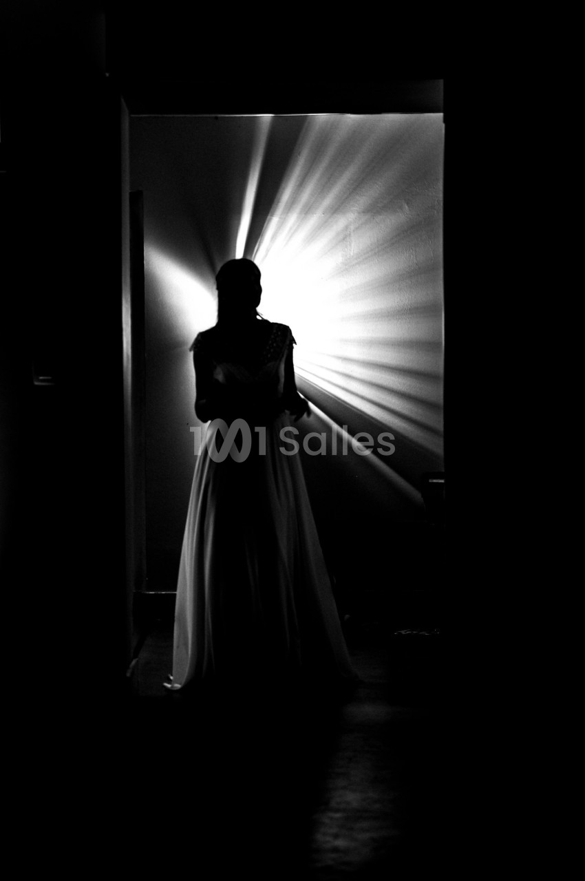 Silhouette d'une femme en robe longue, éclairée par une lumière rayonnante dans un couloir sombre.