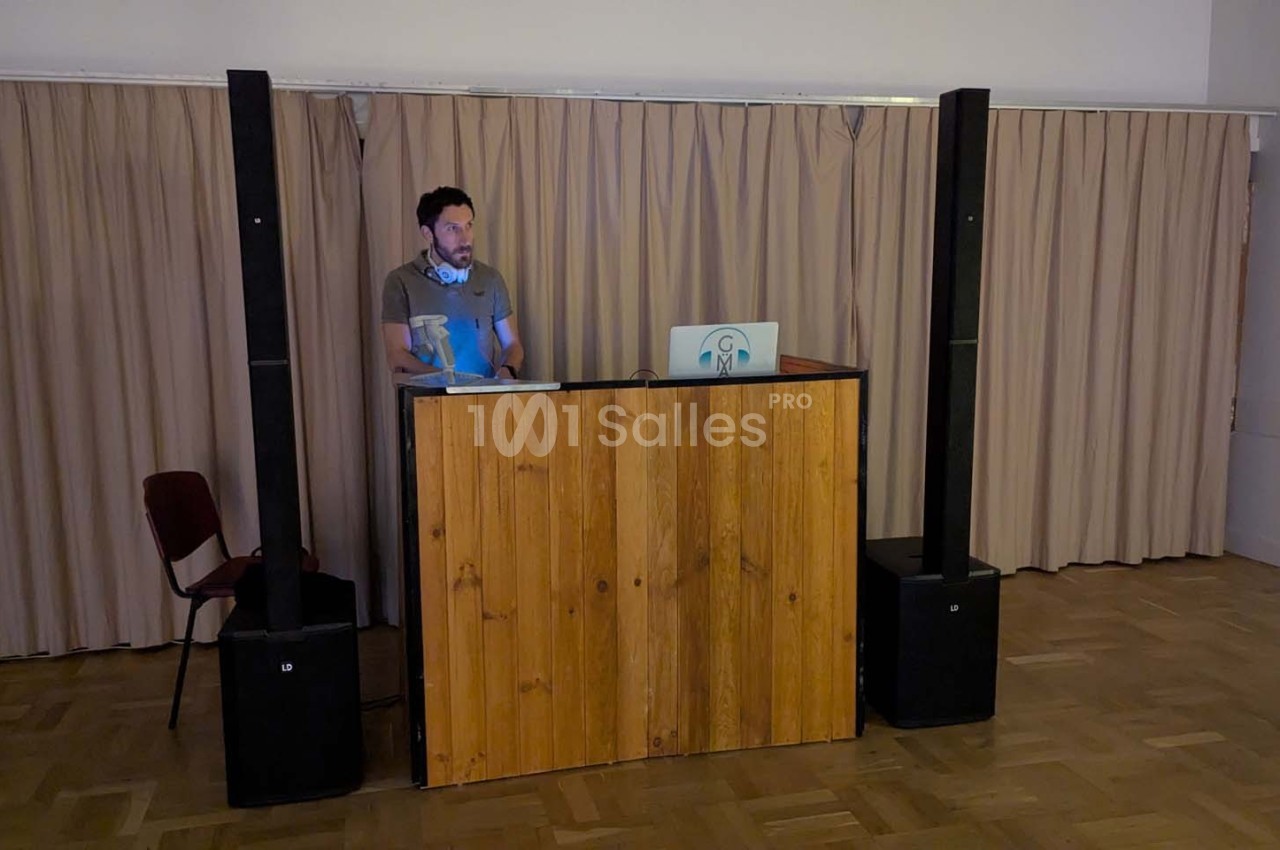 Un homme debout derrière un pupitre en bois avec un ordinateur portable, dans une salle avec rideaux beiges.