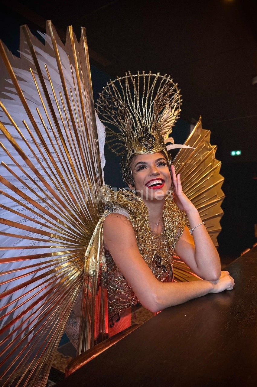 Personne souriante portant un costume doré avec des ailes et une coiffe élaborée, appuyée sur un comptoir.