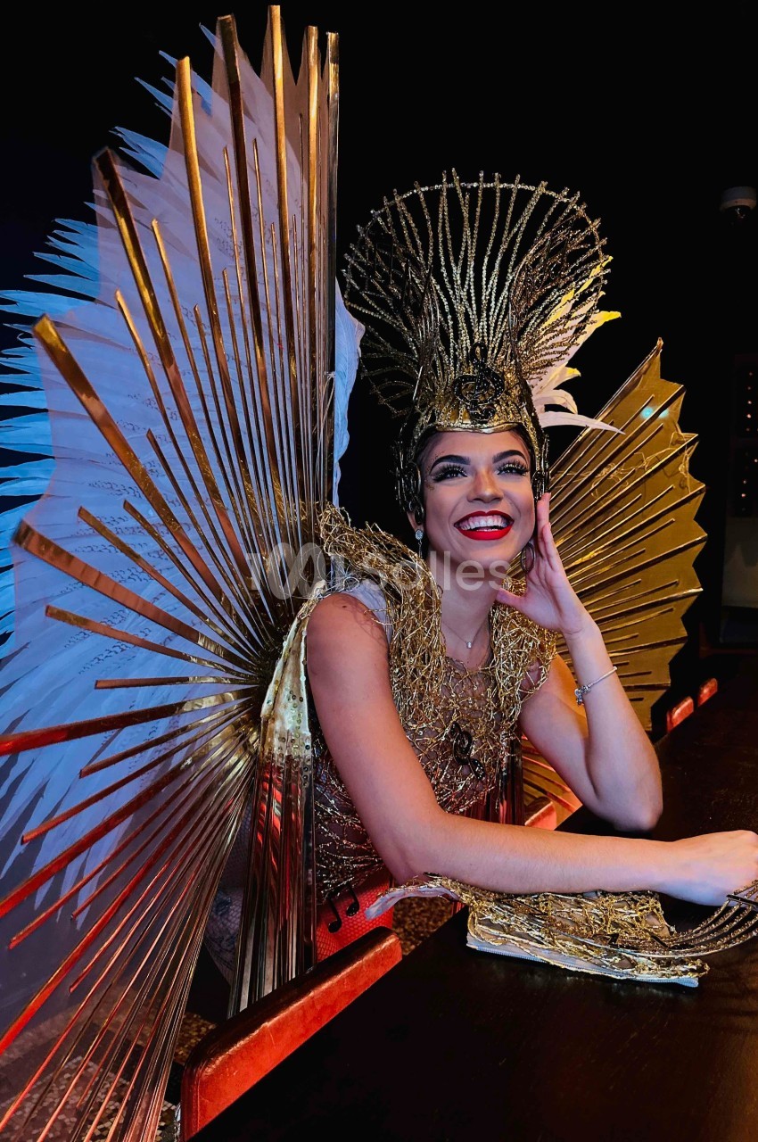Personne souriante portant un costume doré extravagant avec des ailes et une coiffe élaborée, assise à un comptoir.