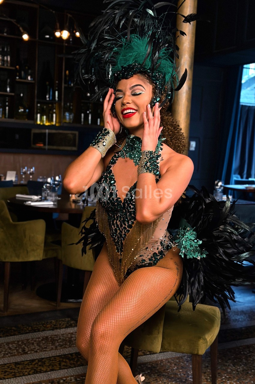 Danseuse en costume à plumes noir et vert, souriante, posant dans un intérieur de restaurant élégant.
