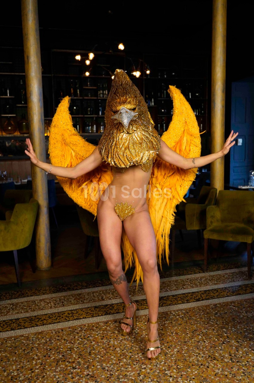 Personne portant un costume doré avec des ailes et un masque en forme de tête d'oiseau, posant dans un intérieur élégant.