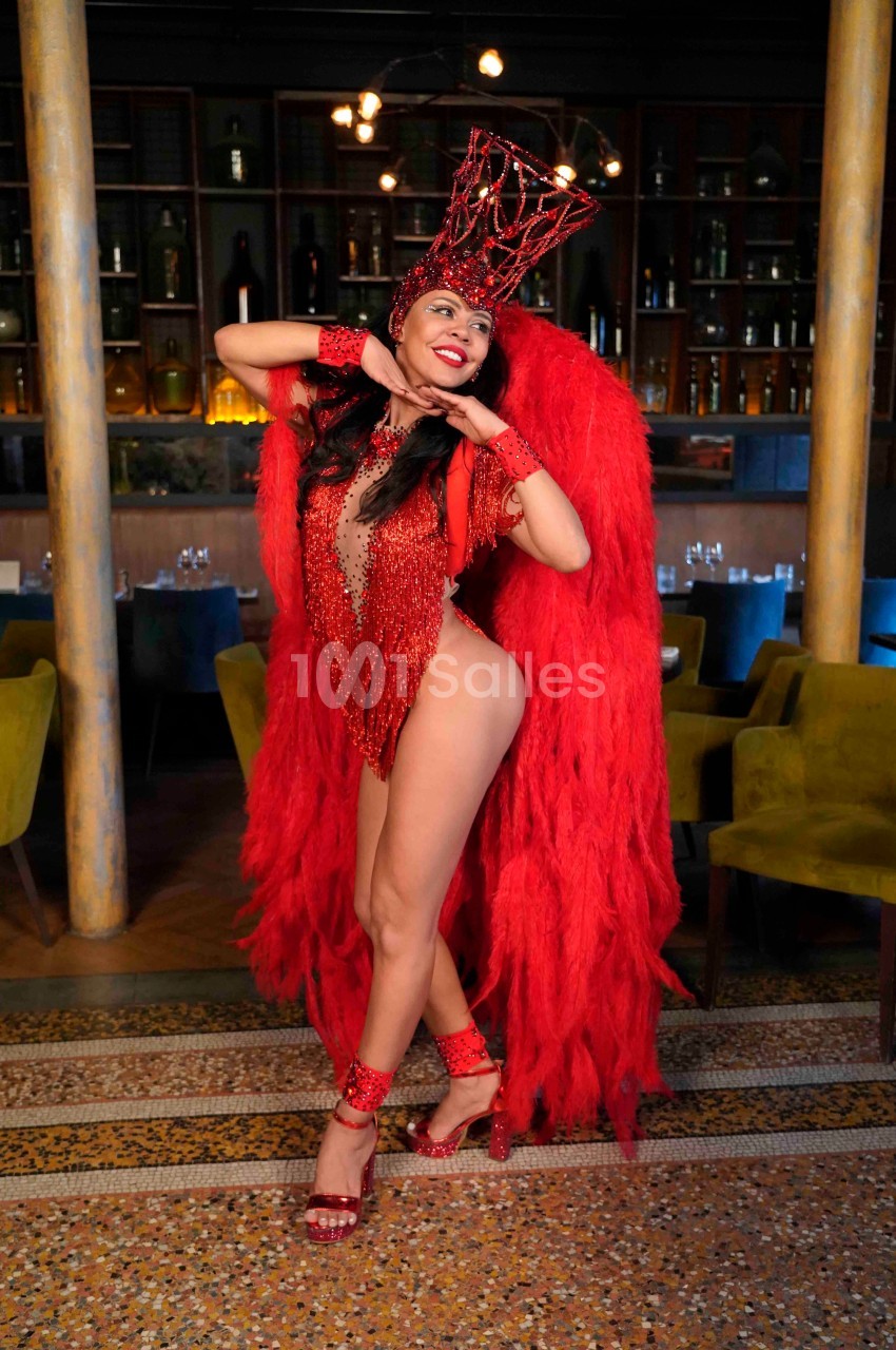 Danseuse en costume rouge à plumes et strass posant dans un intérieur avec tables et étagères en arrière-plan.