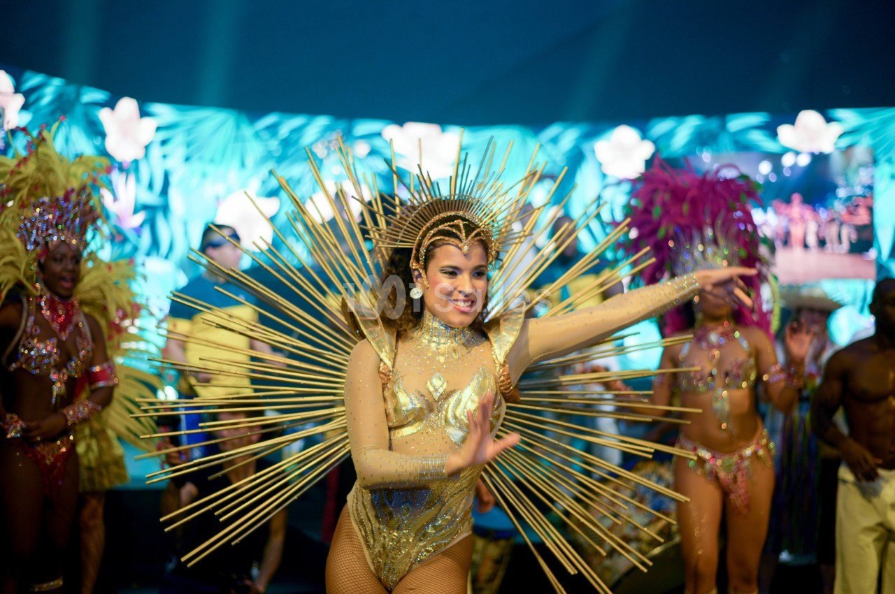 Danseuse en costume doré avec des ornements rayonnants, entourée de performers dans un décor tropical lumineux.