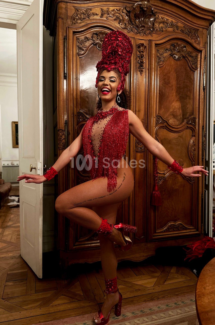Danseuse en tenue rouge ornée de perles et plumes, posant devant une armoire en bois dans une pièce au parquet ancien.