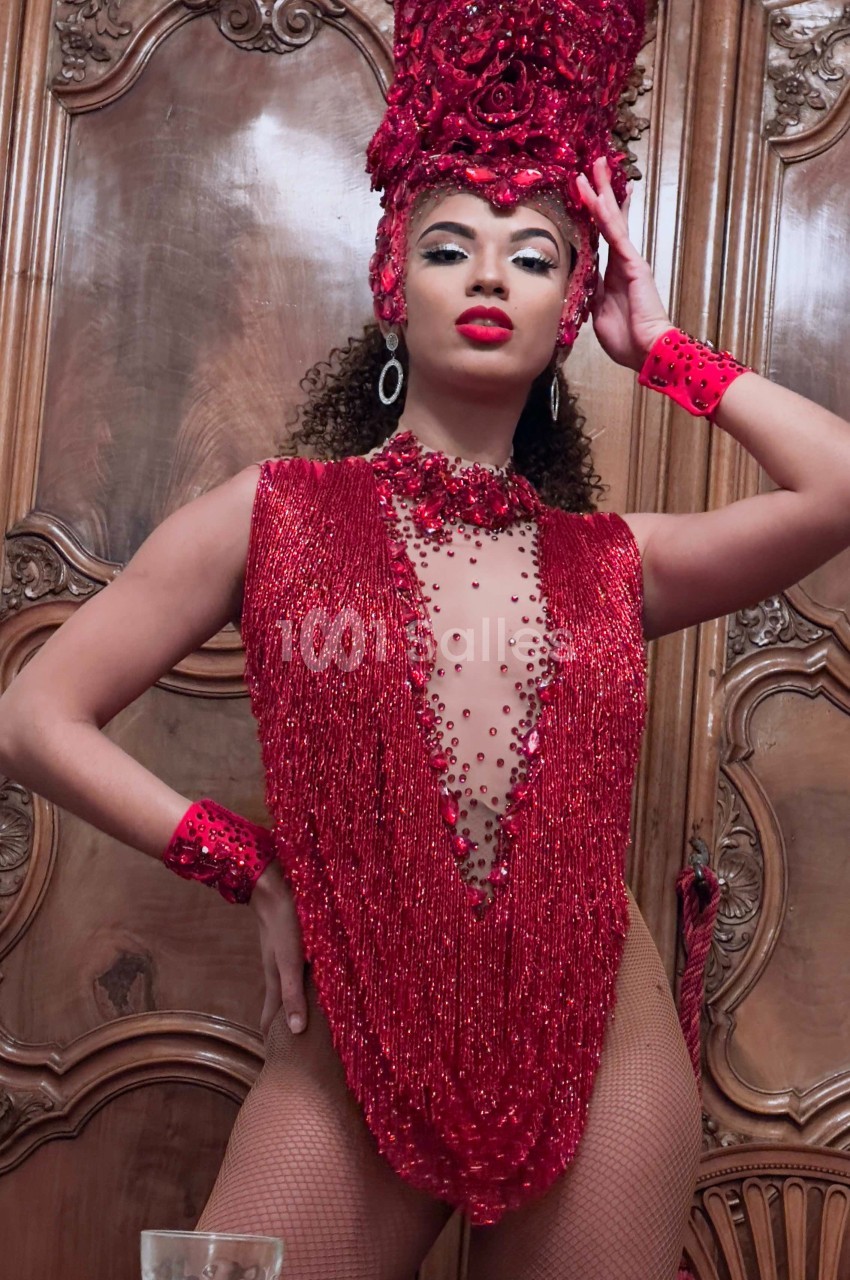 Personne portant une tenue rouge scintillante ornée de perles et un grand accessoire de tête, posant devant un meuble en…