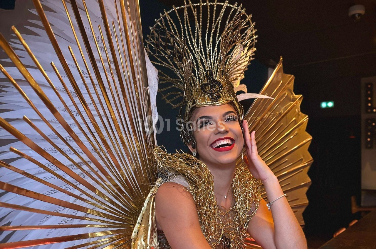 Femme souriante portant une coiffe dorée élaborée et un costume avec des ailes dorées en arrière-plan.