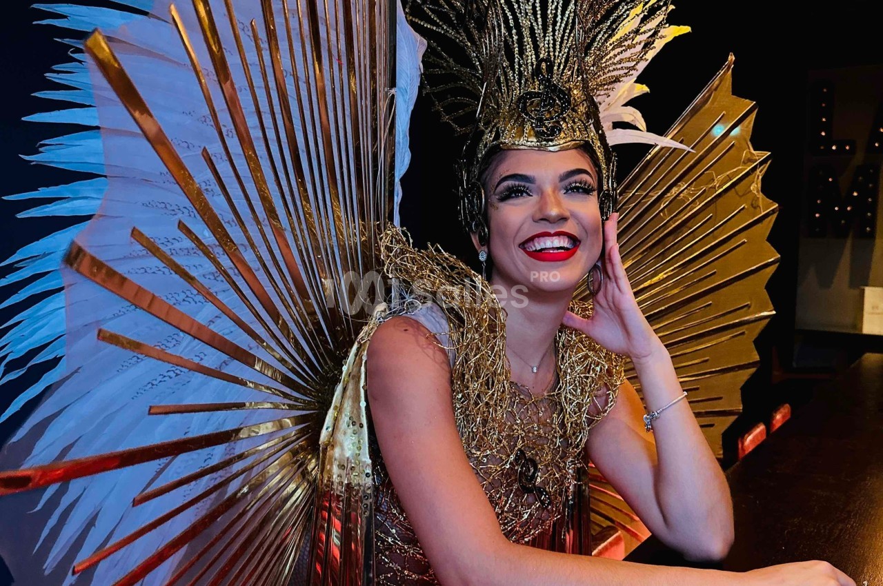 Une femme souriante porte un costume élaboré avec des plumes blanches et des ornements dorés dans un décor sombre.
