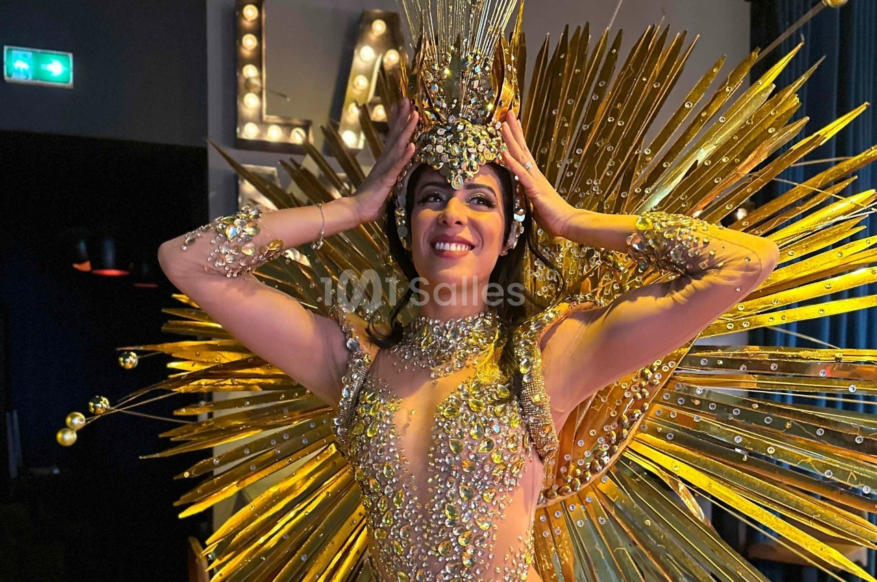 Danseuse en costume doré étincelant avec des ornements scintillants et une coiffe élaborée, souriant sur scène.