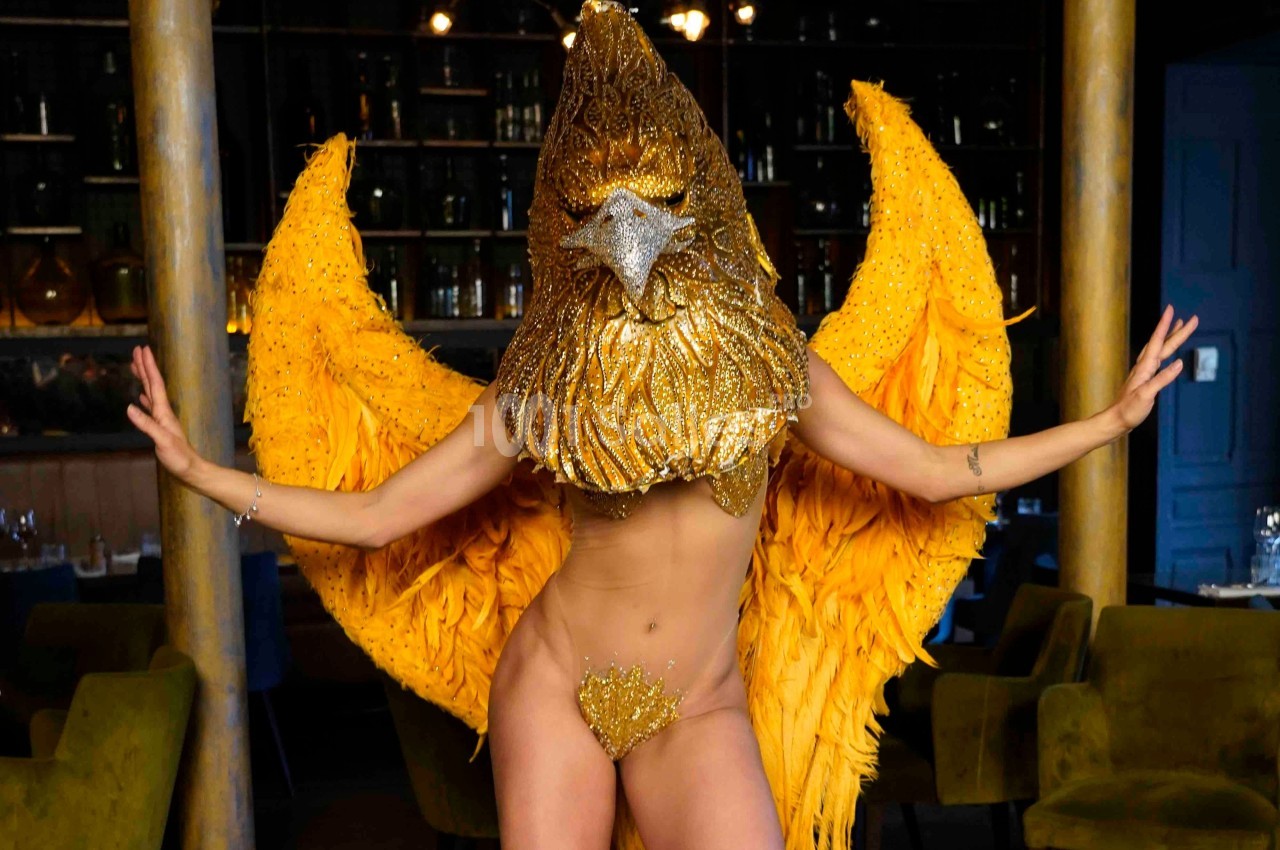 Personne portant un costume doré extravagant avec un masque en forme de tête d'oiseau et des ailes jaunes déployées.