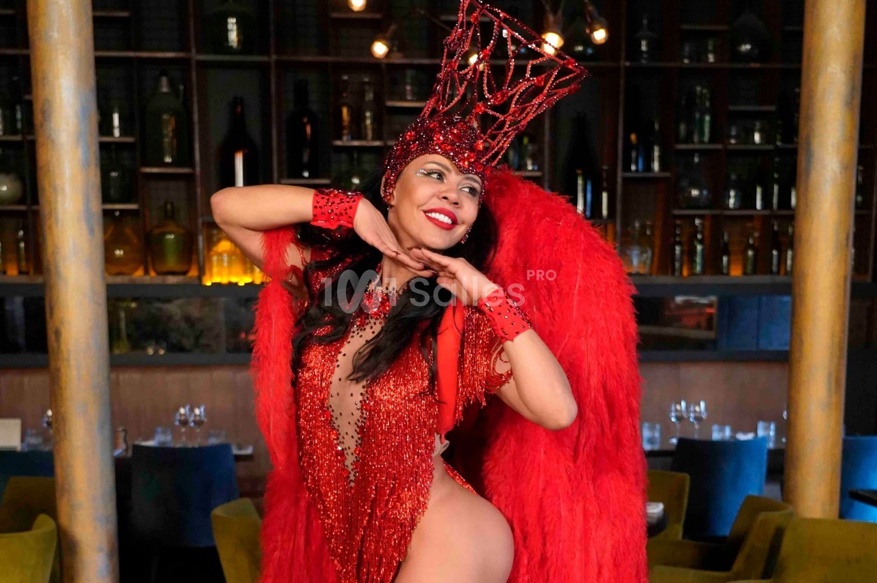 Danseuse en costume rouge à plumes et strass, posant dans un intérieur avec des étagères et des tables en arrière-plan.