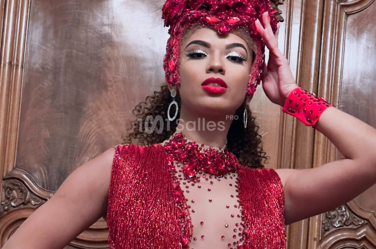 Femme portant une tenue rouge ornée de perles et un accessoire de tête assorti, posant devant une porte en bois sculpté.