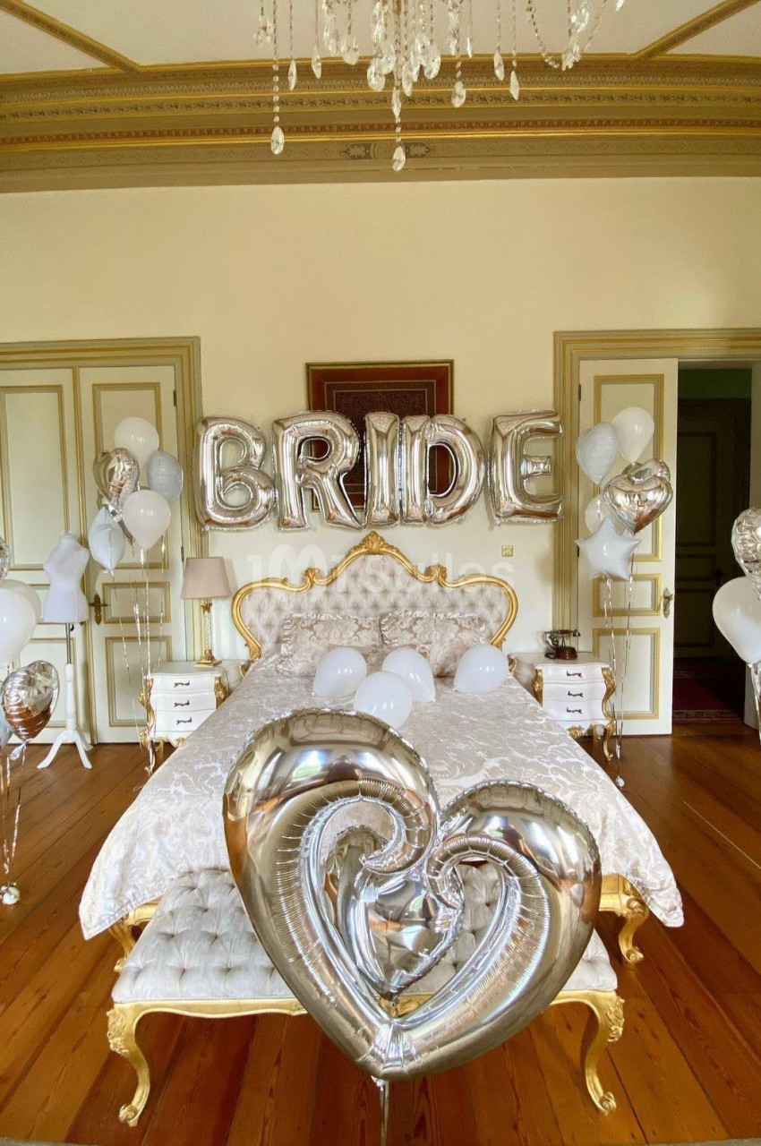 Chambre décorée avec des ballons argentés et blancs, dont des lettres formant ’BRIDE’, au-dessus d'un lit élégant.