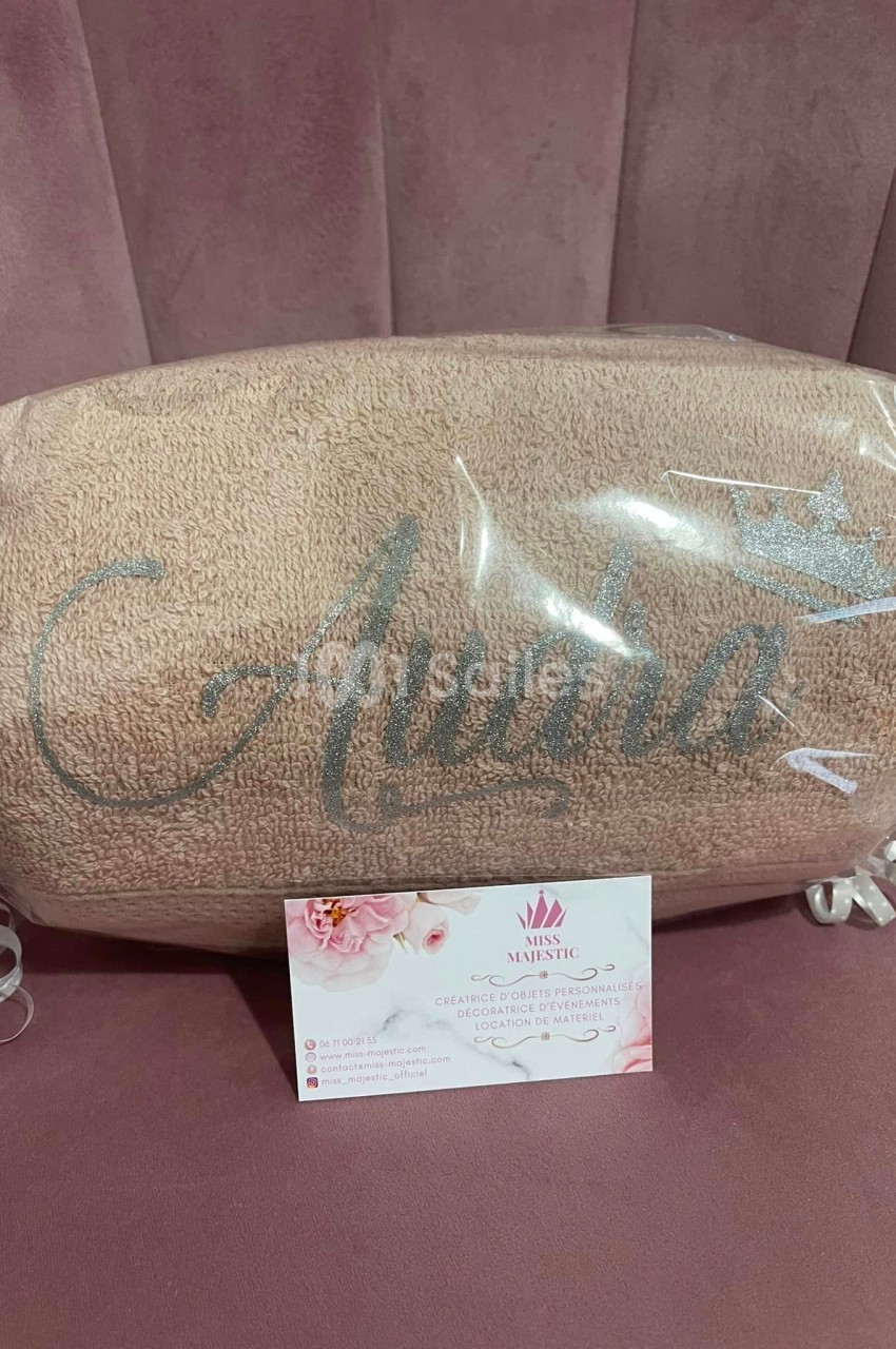 Serviette beige personnalisée avec un prénom brodé et une couronne, emballée dans un plastique transparent sur un fond rose.