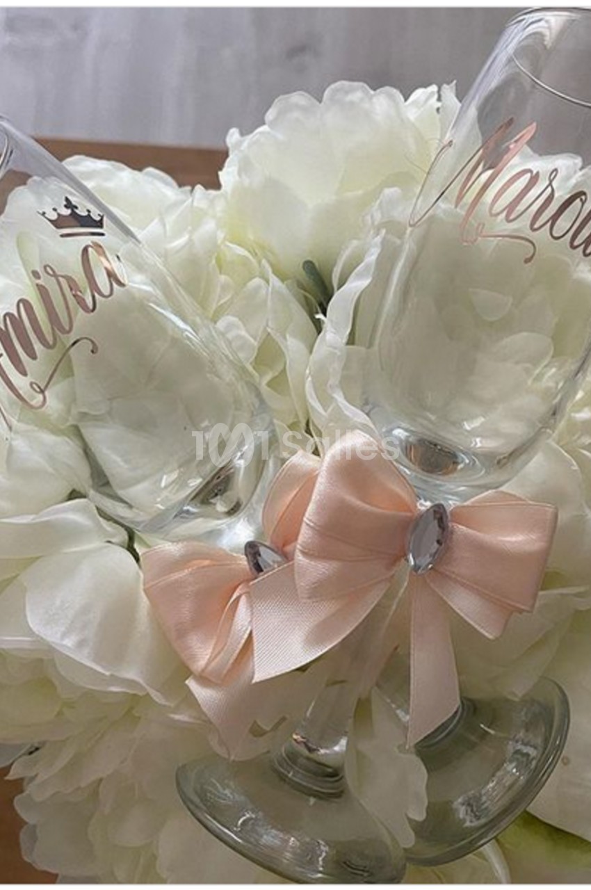 Deux flûtes gravées avec des prénoms reposent sur un bouquet de fleurs blanches, ornées d'un nœud rose pâle.