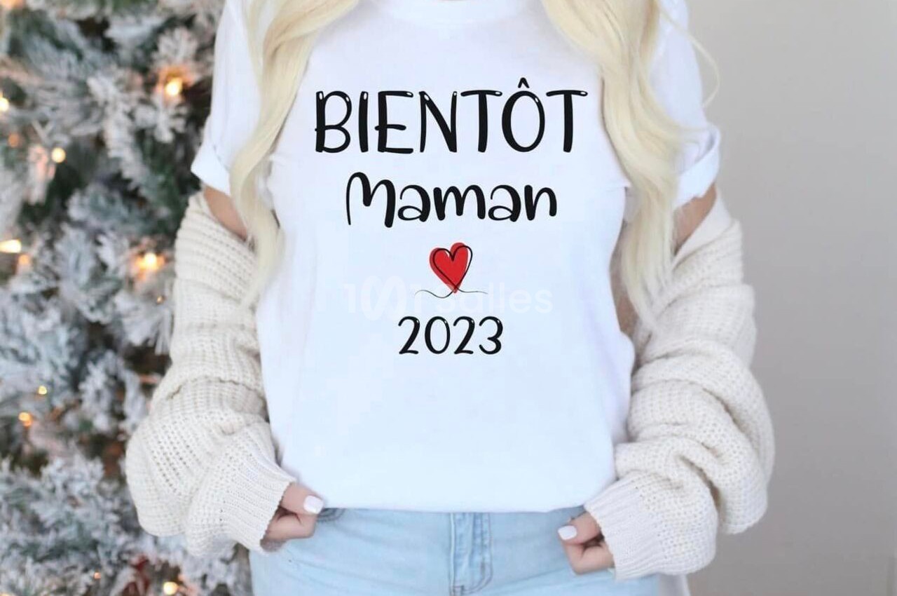 Femme portant un t-shirt blanc avec l'inscription ’Bientôt maman 2023’ devant un sapin enneigé.