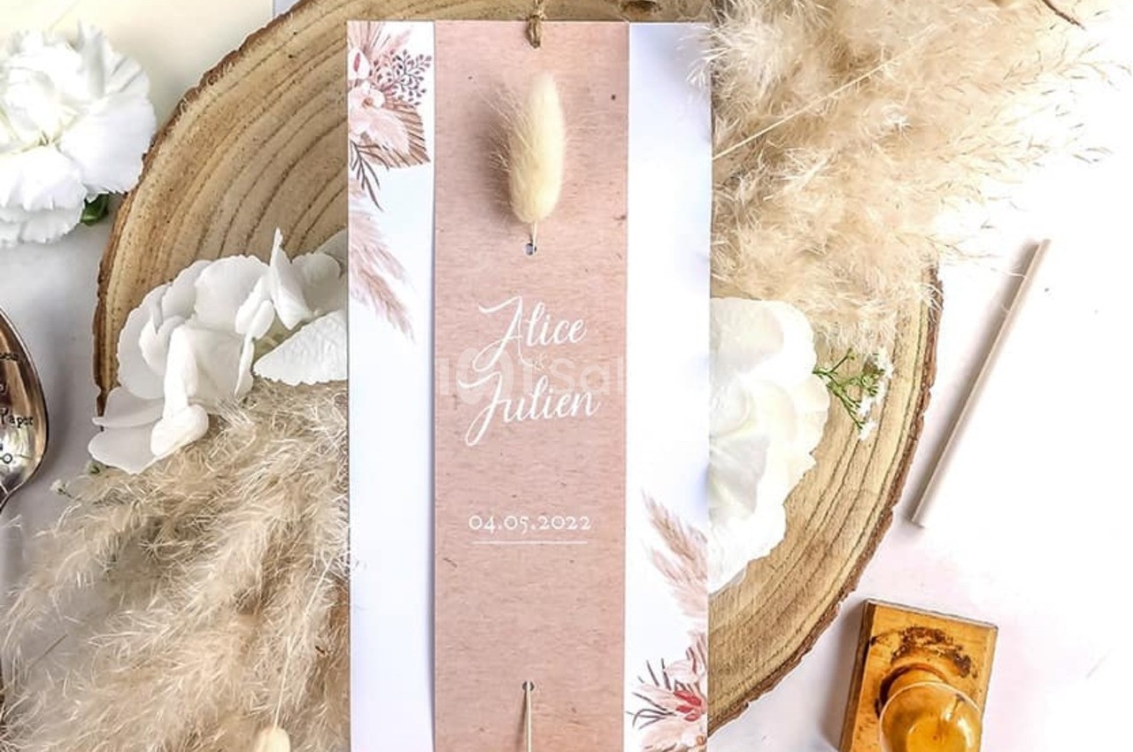 Faire-part de mariage posé sur une tranche de bois, entouré de fleurs blanches, pampas et cachet de cire.