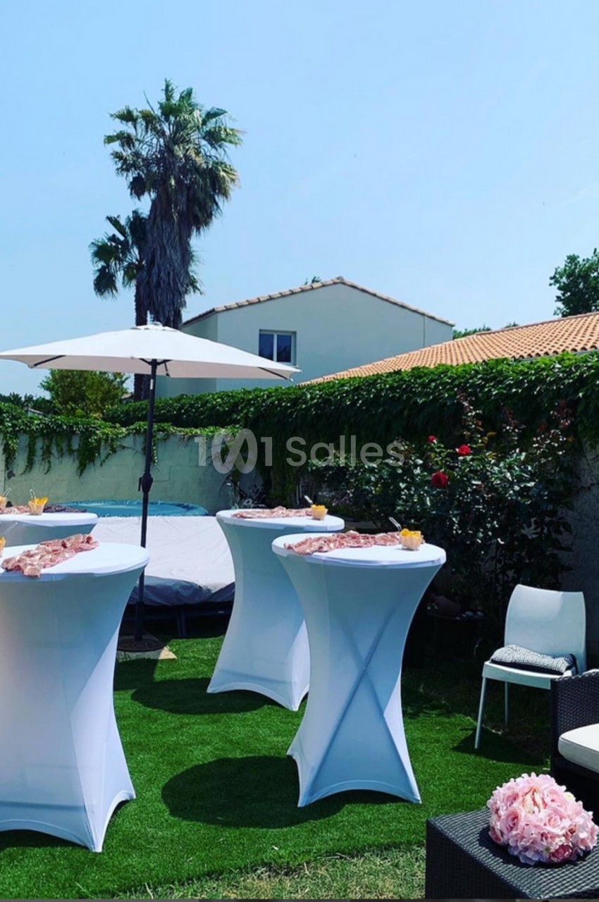 Tables hautes dressées avec des amuse-bouches dans un jardin ensoleillé, entourées de chaises et d'un parasol.