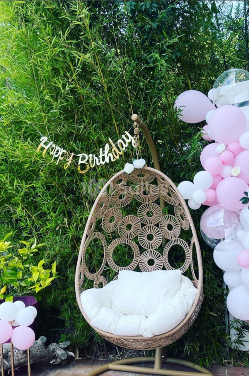 Fauteuil suspendu en rotin avec coussin blanc, entouré de ballons roses et blancs, devant un fond de bambous.