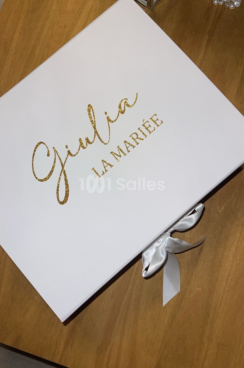 Boîte blanche avec l'inscription dorée ’Giulia La Mariée’ posée sur une table en bois, fermée par un ruban blanc.