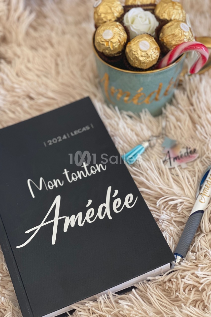 Carnet noir ’Mon tonton Amédée 2024’ posé sur un plaid beige, avec une tasse contenant des chocolats et un stylo.