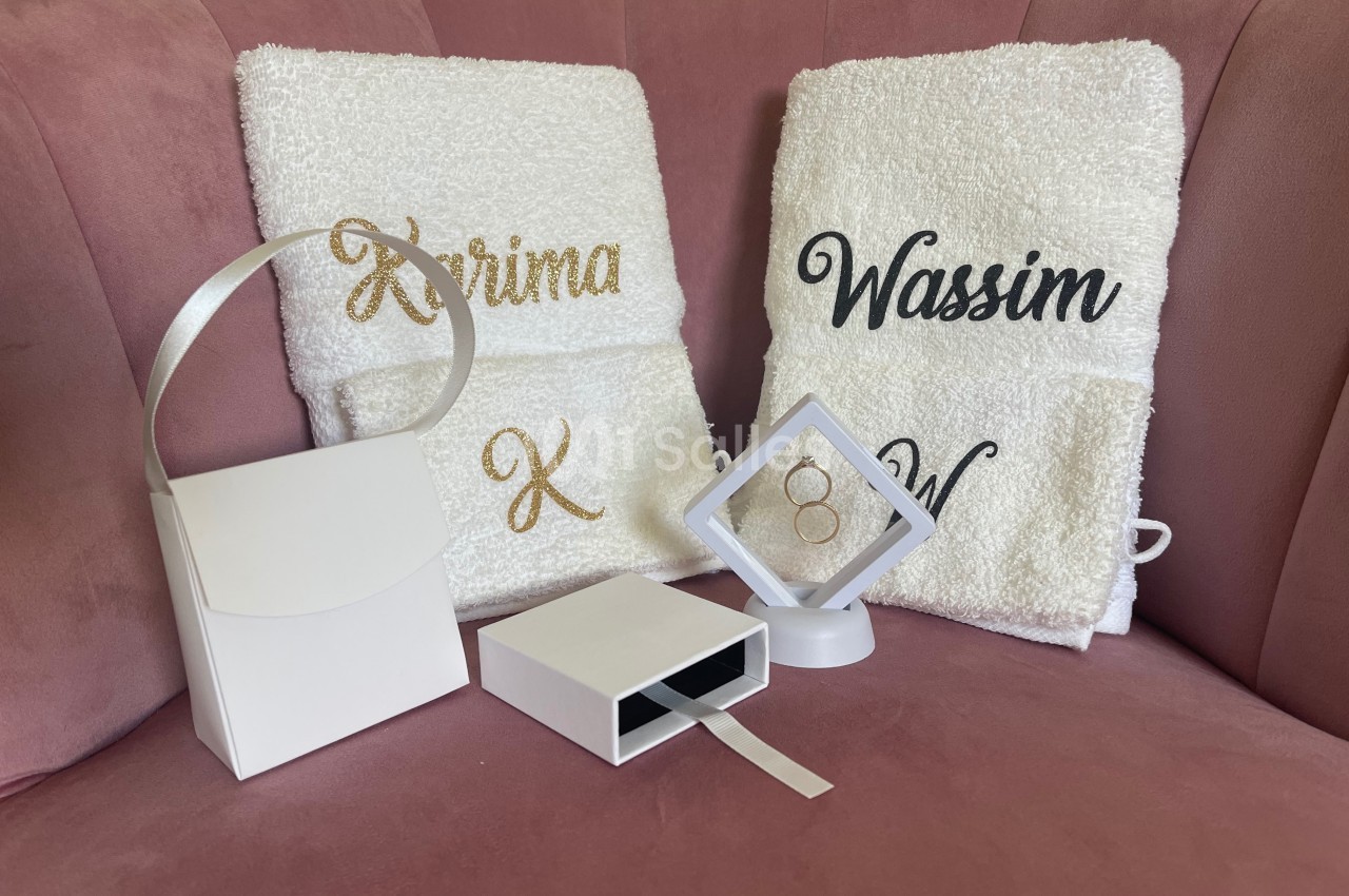 Serviettes personnalisées avec des prénoms brodés, posées sur un fauteuil rose, accompagnées de boîtes blanches.
