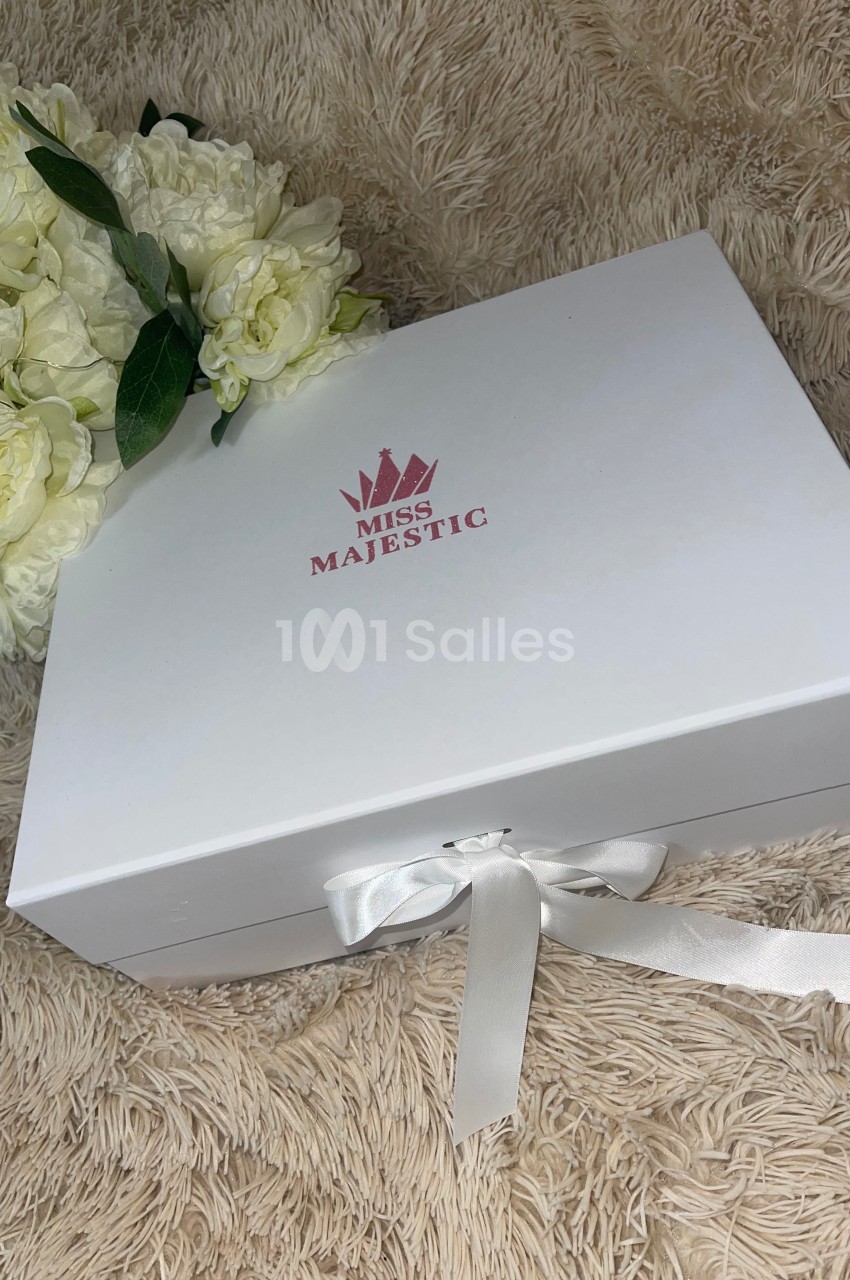 Boîte blanche avec un ruban, logo ’Miss Majestic’ rose, posée sur un tapis beige près d'un bouquet de fleurs blanches.