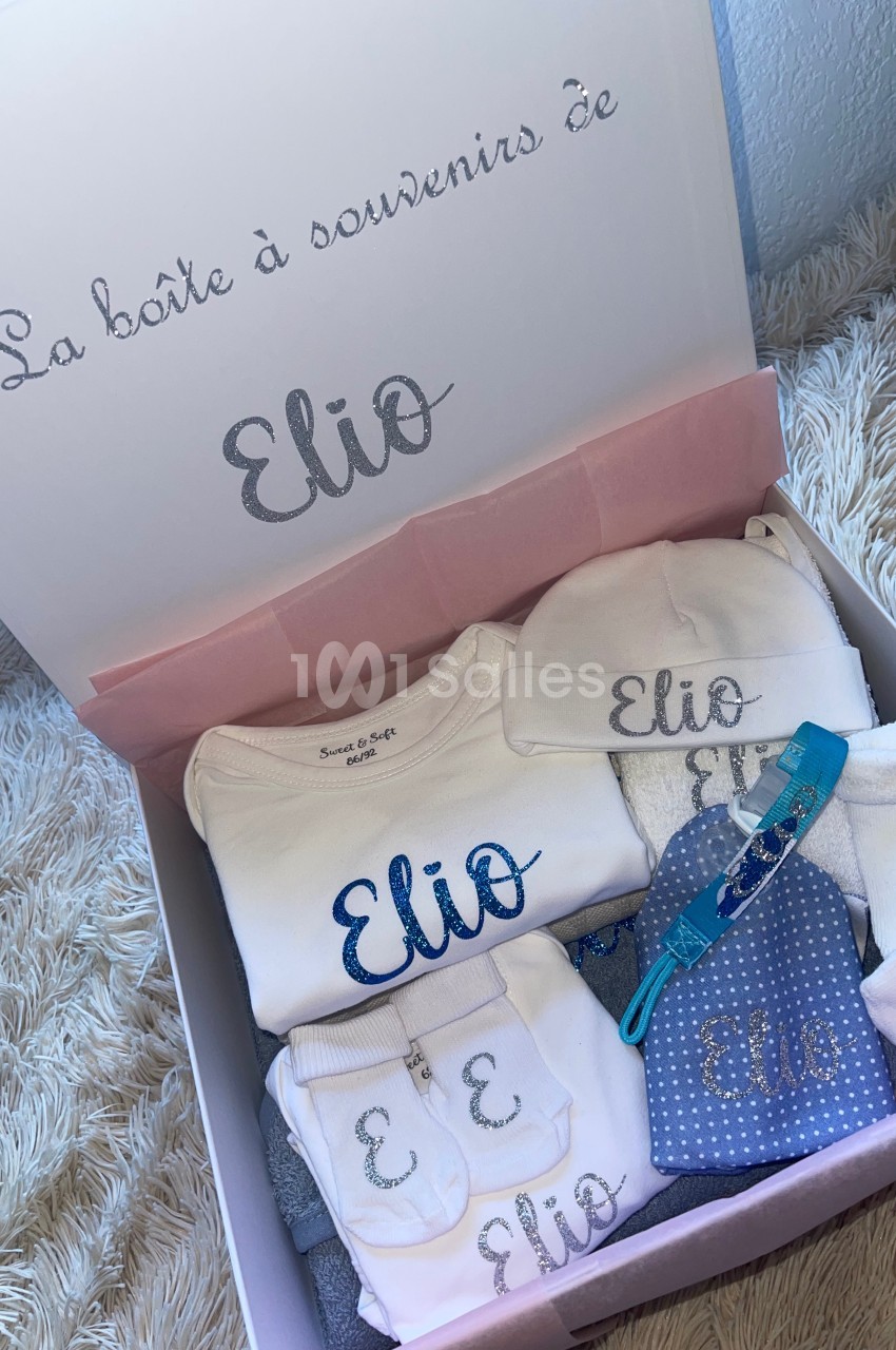 Boîte contenant des vêtements et accessoires pour bébé personnalisés avec le prénom ’Elio’, présentés sur un fond doux.