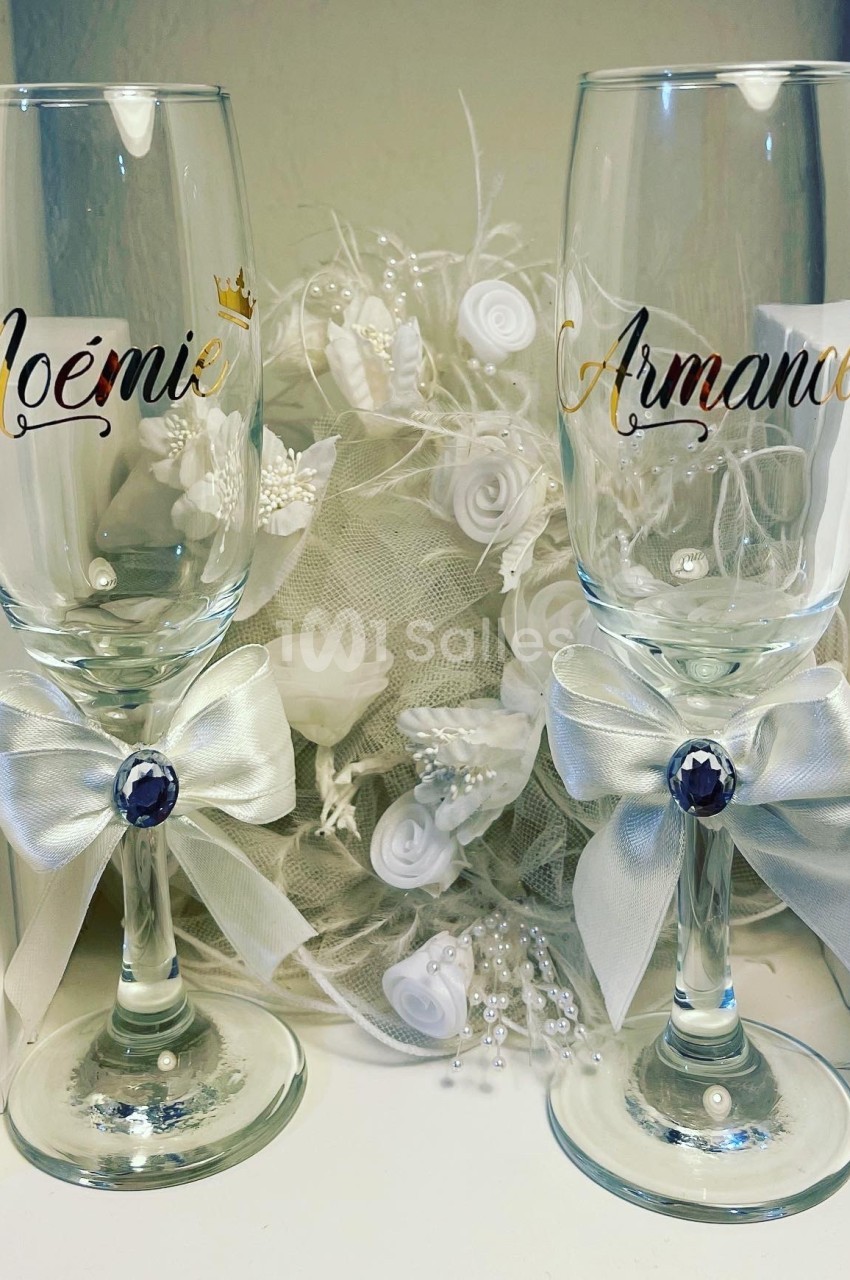 Deux flûtes à champagne personnalisées avec des prénoms, ornées de nœuds blancs et posées devant une décoration florale.