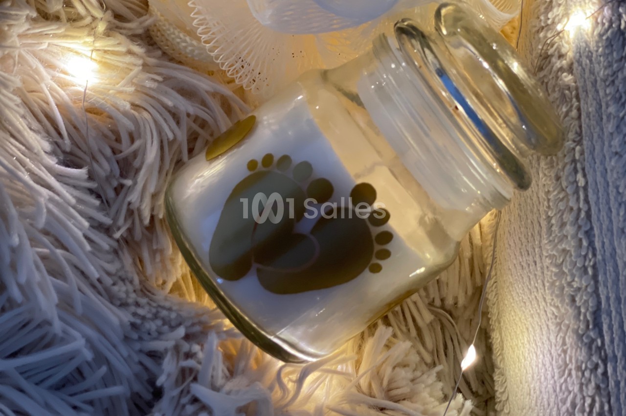 Bougie dans un pot en verre décoré d'empreintes de pieds, posée sur un tapis texturé avec des lumières douces.