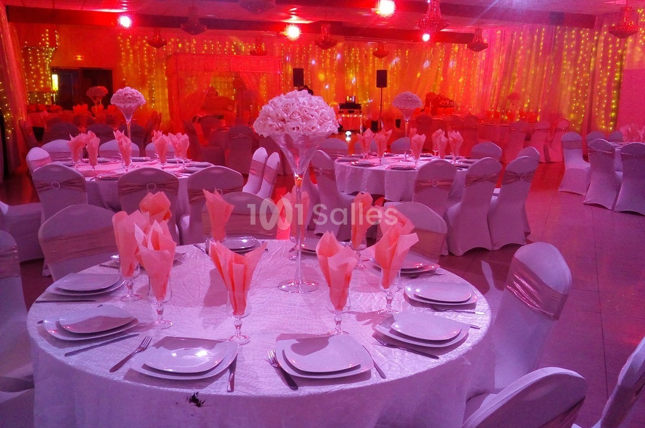 Salle de réception décorée avec des tables rondes dressées, nappes blanches, serviettes roses et éclairage rouge tamisé.