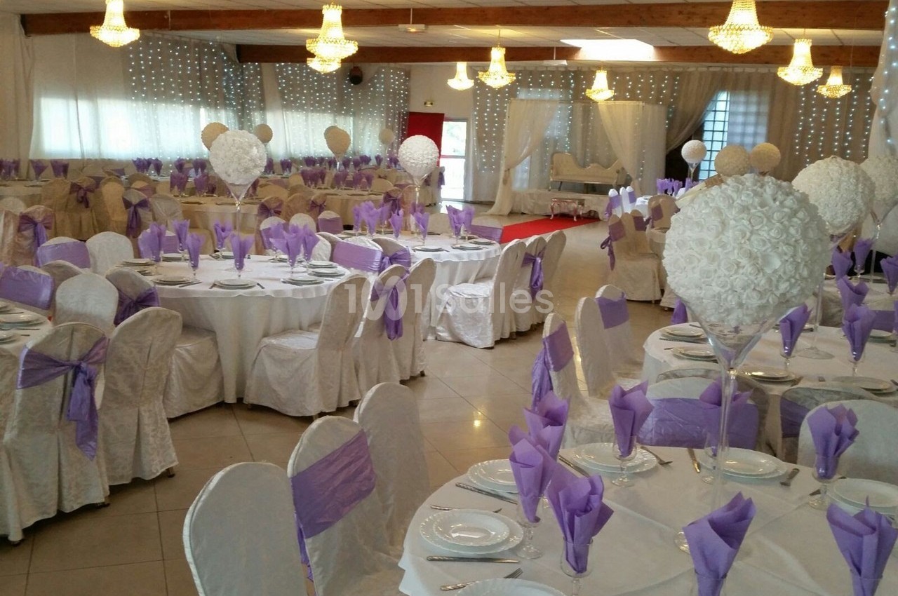 Salle de réception décorée avec des tables rondes, nappes blanches, serviettes violettes et centres de table fleuris.