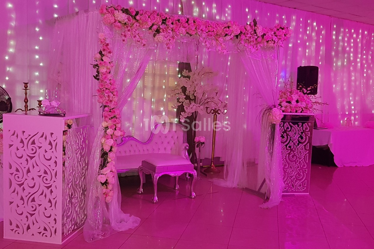 Décor de mariage avec arche florale, canapé blanc, rideaux lumineux et éclairage rose dans une salle de réception.