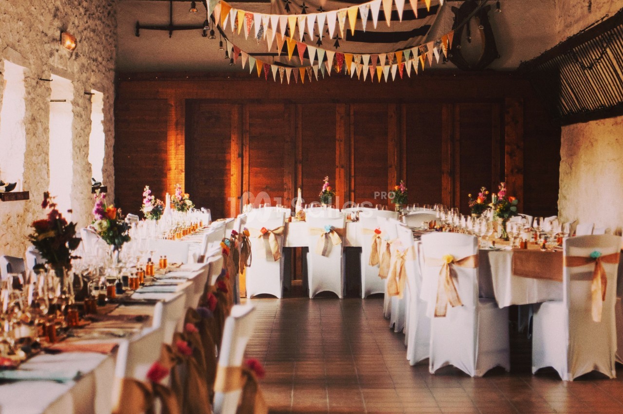 Salle décorée pour un événement avec longues tables dressées, chaises ornées de rubans et guirlandes suspendues.