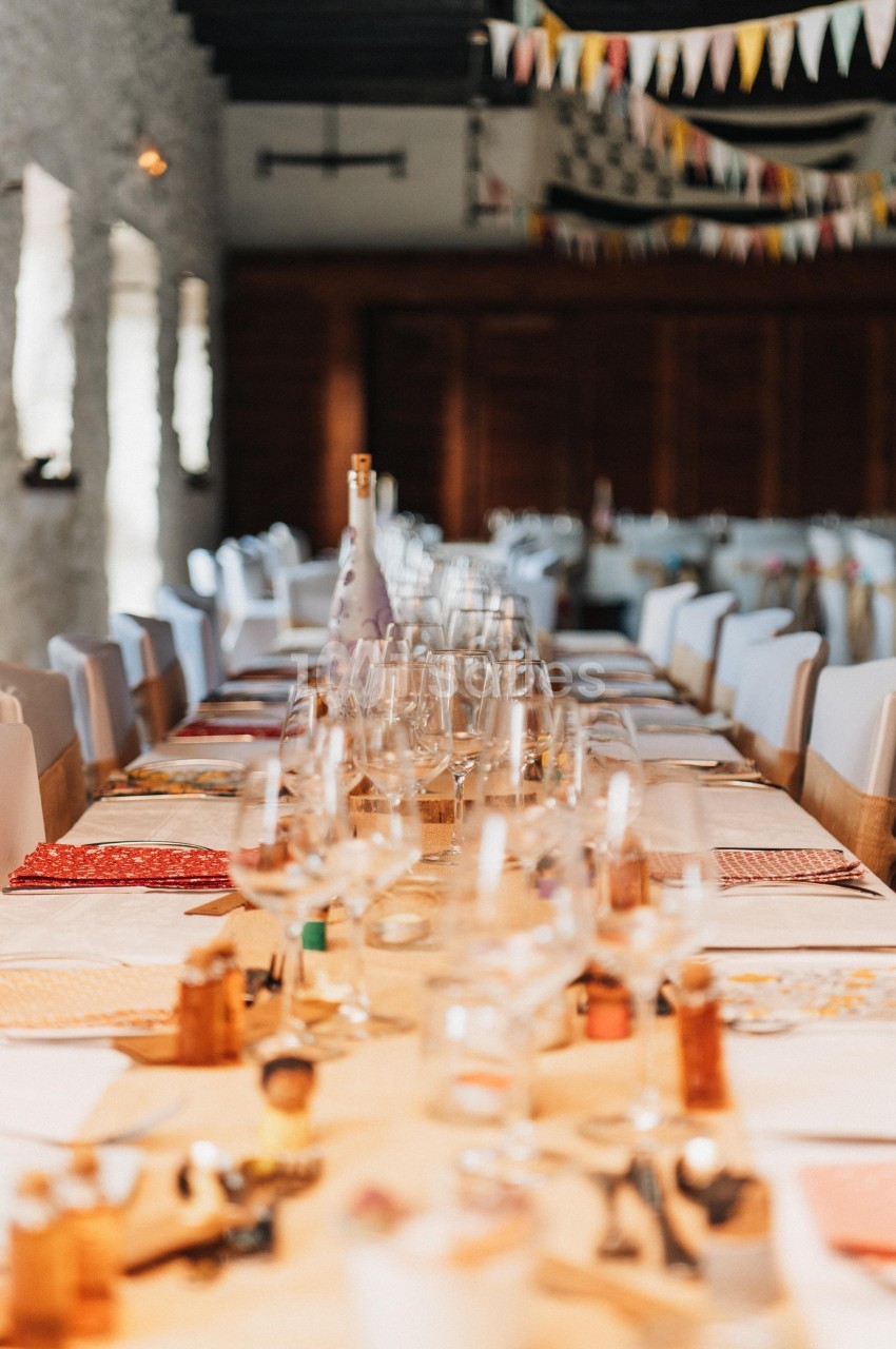 Table décorée pour un repas festif, avec nappes colorées, verres et guirlandes suspendues dans une salle lumineuse.